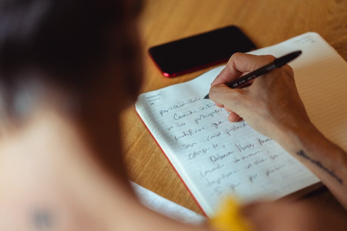Person writing notes in a paper planner beside a face-down smartphone.